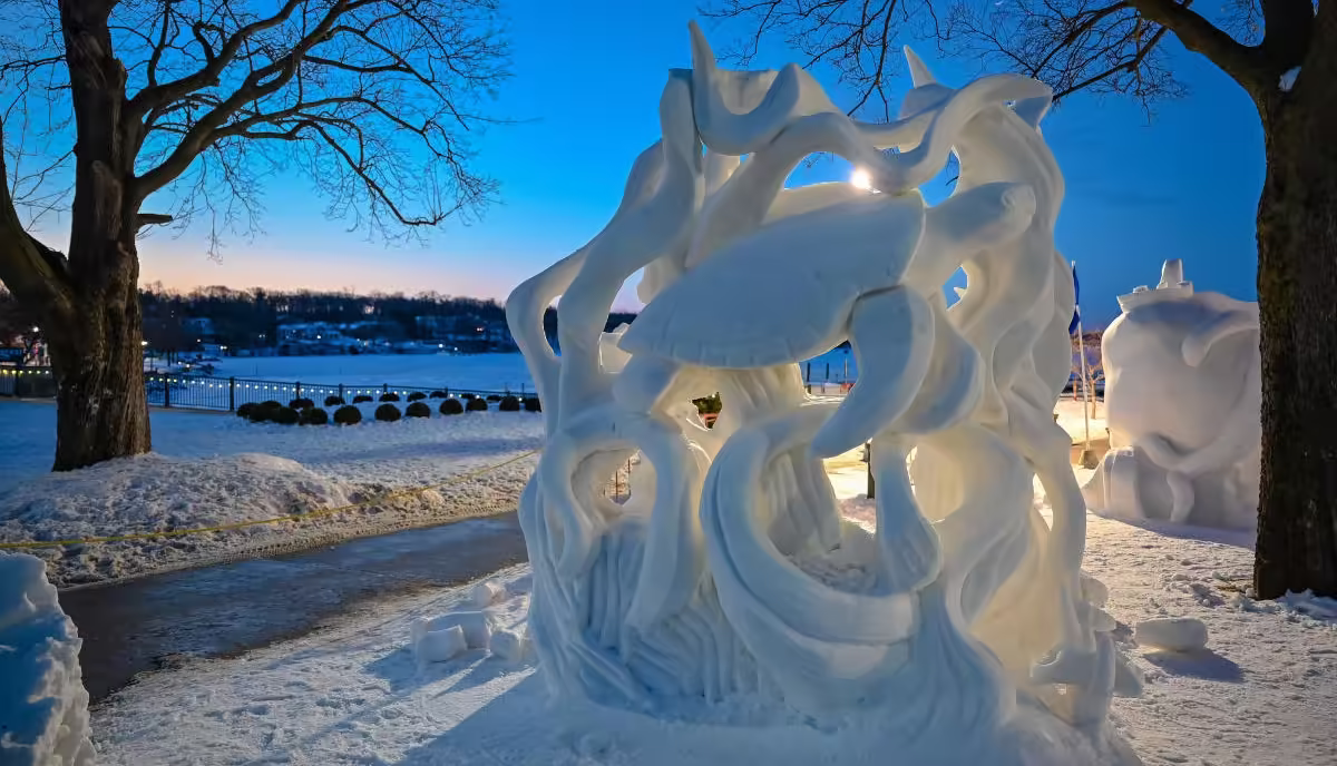 Lake Geneva Ice sculpture displayed in a snowy park near a frozen lake at dusk, with trees and winter lights in the background